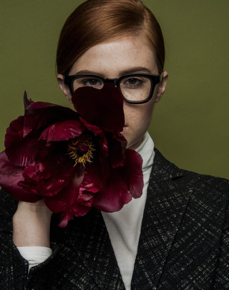 A red-headed woman in a suit jacket with black, plastic glasses and a white shirt, holding red flowers