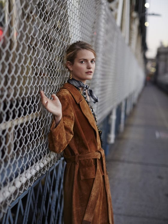 A woman wearing a long suede jacket leaning against a wire gate while looking outward to the camera