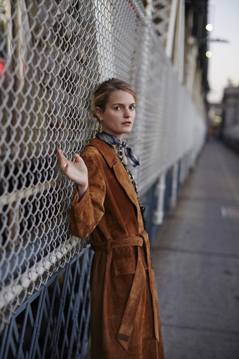A woman wearing a long suede jacket leaning against a wire gate while looking outward to the camera