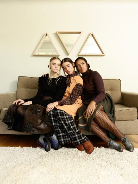 Three women- Caucasian, Latina and Black- seated on a couch with three triangular mirrors frames on the wall behind them