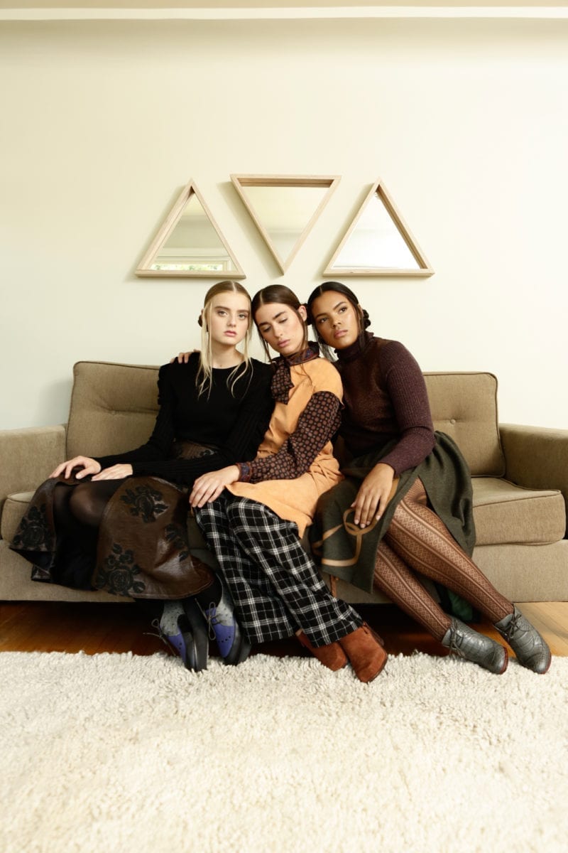 Three women- Caucasian, Latina and Black- seated on a couch with three triangular mirrors frames on the wall behind them
