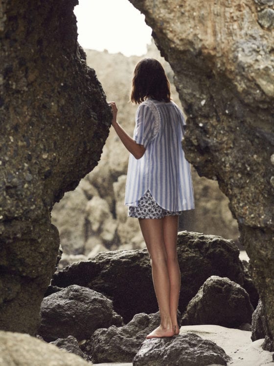 Woman in striped shirt and shorts standing in a alcove of large rocks lookin out in the distance