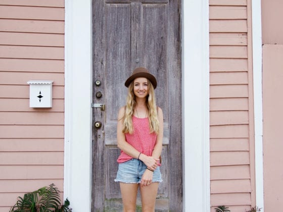 A woman in shorts, a tank top and fedora hat standing in front of the door of a home