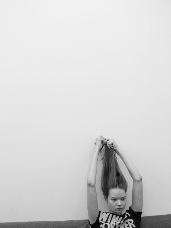 A black and white photo of a girl pulling her hair as she sits against a wall
