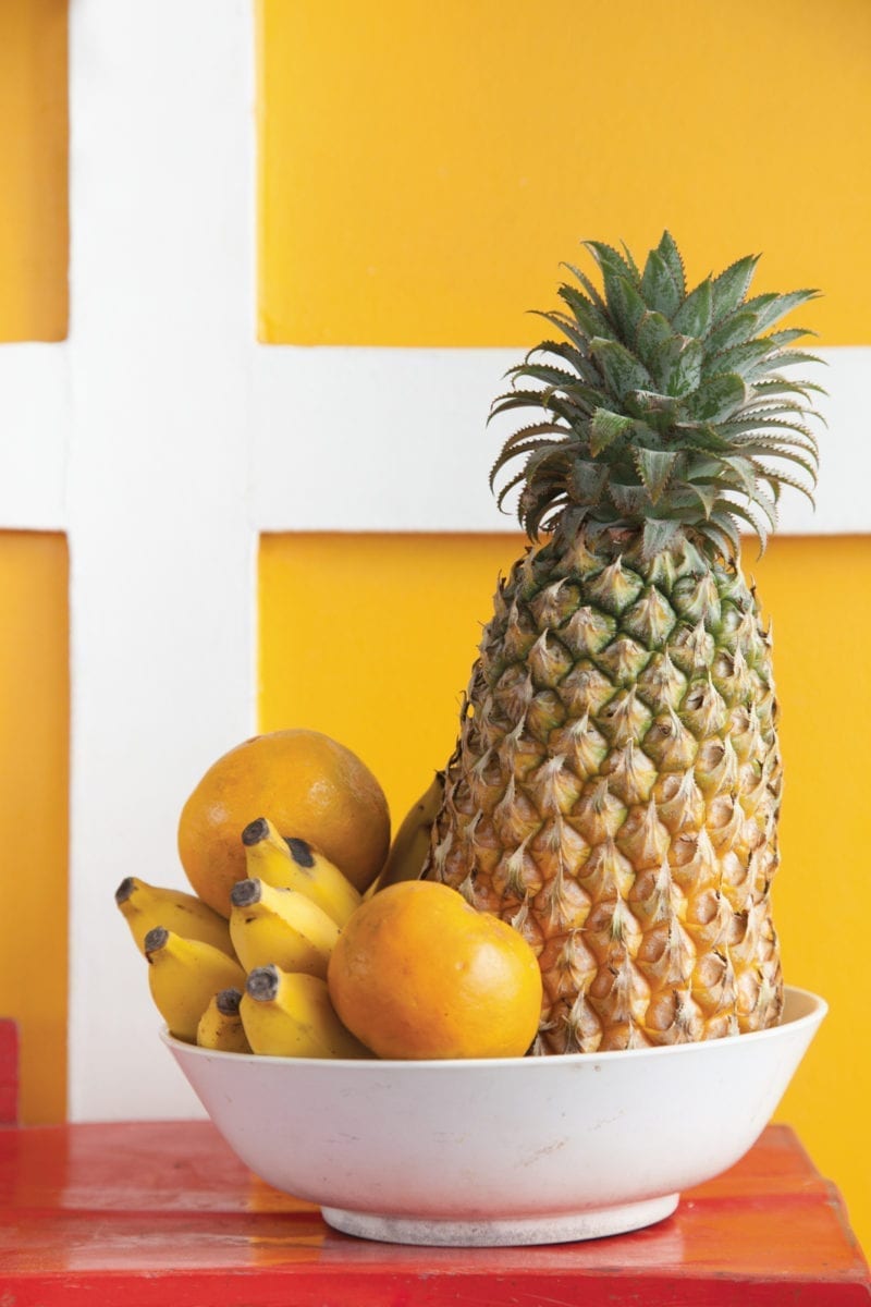 A photo of a fruit bowl with pineapple, banana and melon in front of a yellow wall