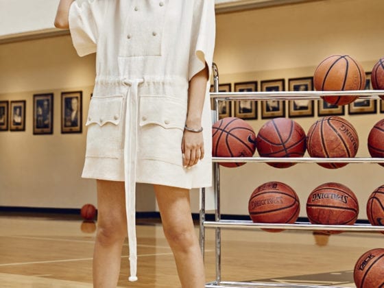 A woman standing in a gym wearing athleisure clothes, holding a basketball near her head as she stands in front of a rack of basketballs