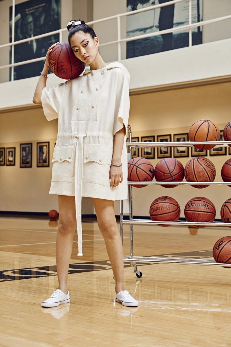 A woman standing in a gym wearing athleisure clothes, holding a basketball near her head as she stands in front of a rack of basketballs