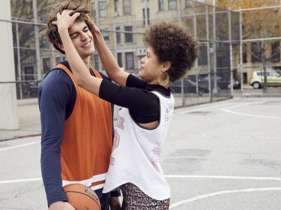 A man and woman standing on a basketball court as she plays with his hair and he smiles