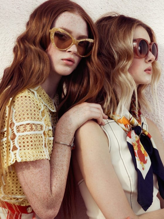 Two girls wearing sunglasses and one leans over the other's shoulder