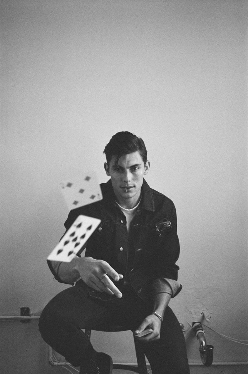 A man seated on a stool as he throws cards toward the camera lens