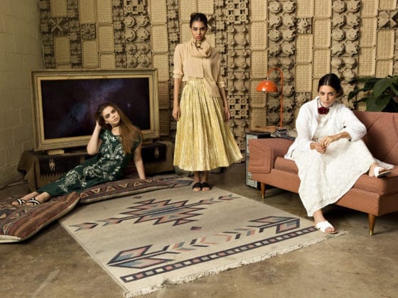 A group of three woman in a small living room, one seated on the couch, another on the floor and one standing