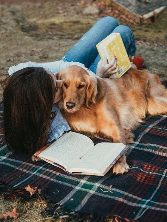 A woman lying on a blanket next to her dog as she reads