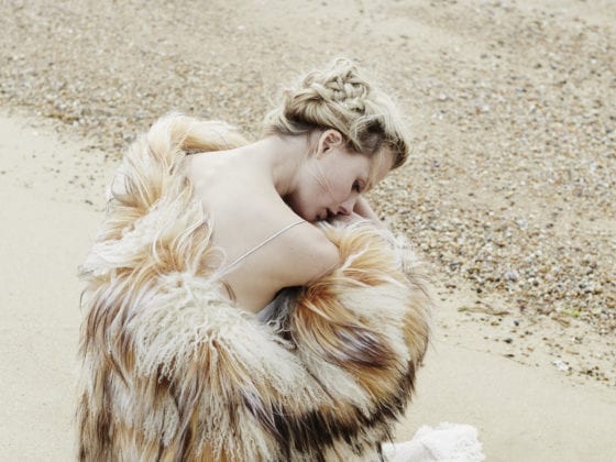 A woman with fur jacket falling off her shoulder as she crouches in the sand near a beach