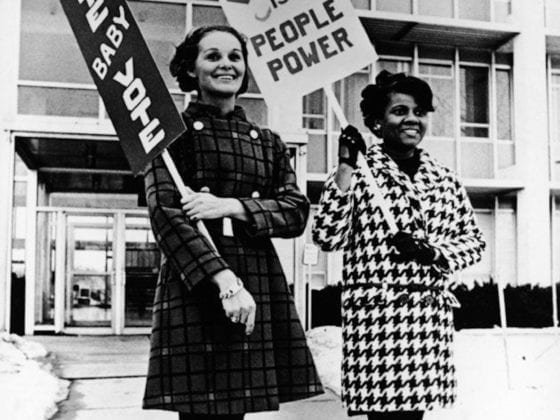 Two women protesting with signs that say, "Vote baby vote," and "Voting is people power."