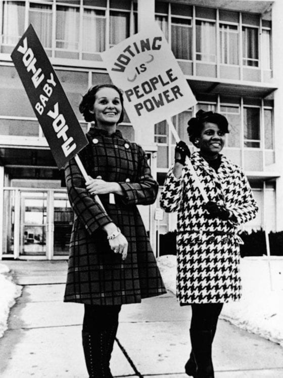 Two women protesting with signs that say, "Vote baby vote," and "Voting is people power."