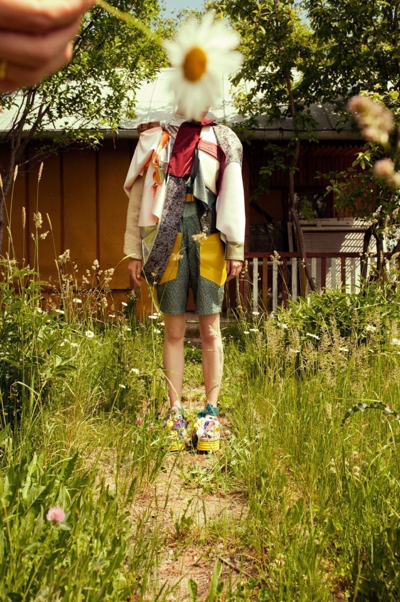 A woman standing in an overgrown yard as hand in the foreground holds a flower that obscures her face