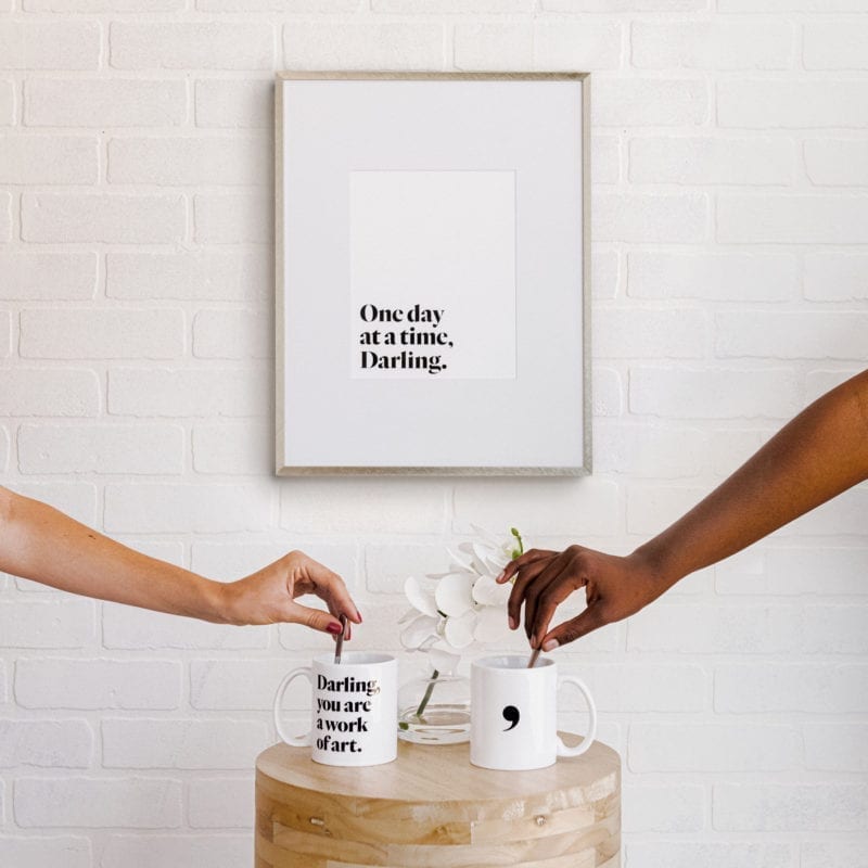 Two hands stirring coffee cups with a piece of art on the wall that says, "One day at a time, Darling."