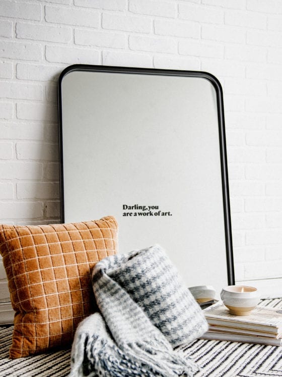 A mirror leaning against a wall with a pillow, throw blanket and a pile of books on the floor