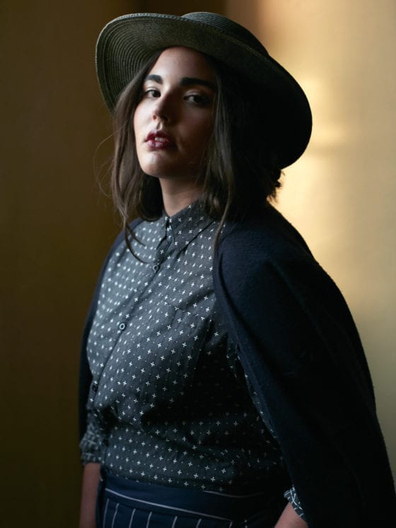 A woman in a shadowy hall wearing a fedora and black polka dotted dress