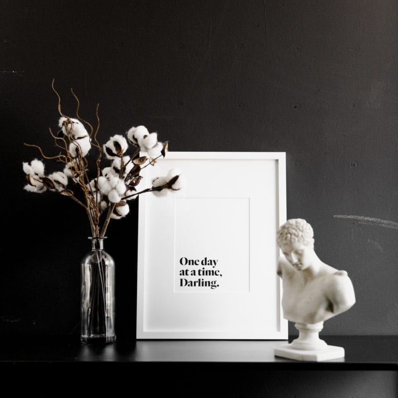 A print that says "One day at a time, Darling" next to a vase of flowers and a sculpture