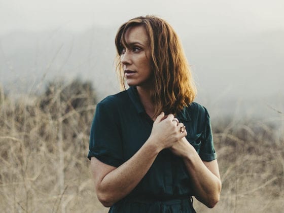 A woman in a field tugging on her hair as she looks over her shoulder to the right