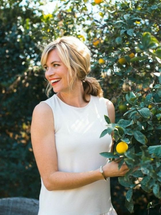 A smiling woman looking over her shoulders as she stands next to a lemon tree